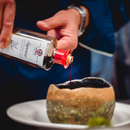 A person pours Taycte Acetaia Malpighi Saporoso Balsamic Vinegar of Modena — a rich and savory Italian condiment — from a glass bottle with a red cap into a rustic, green-and-brown cup resting on a white plate.