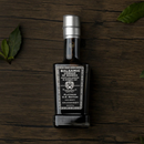 A bottle of Taycte Acetaia Malpighi Platinum IGP – High-Density Balsamic Vinegar of Modena, aged over 25 years, crafted by the renowned Malpighi family, rests on a dark wooden surface amidst a few green leaves. The label bears the words “Platinum U.S. Edition” in white text, hinting at the extra-aged balsamic vinegar inside.