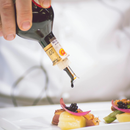 A hand pours Taycte Acetaia Malpighi D.O.P. Traditional Balsamic Vinegar of Modena — Affinato Grade, aged over 12 years, over a gourmet dish elegantly garnished with onions, caviar, and cheese on a white plate.