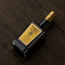 A bottle of Taycte Acetaia Malpighi Gold Balsamic Vinegar of Modena — IGP Certified, Aged 15-20 Years sits on a dark wooden surface with its gold label gleaming.