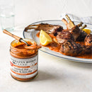 A jar of Terrapin Ridge Farms Roasted Red Pepper & Feta Dip with a spoon sits beside grilled lamb chops and lemon wedges—a perfect Mediterranean appetizer. A glass of water completes this delicious Terrapin Ridge Farms dip spread.