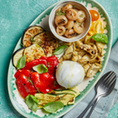 An oval plate showcasing a colorful Italian antipasto features grilled zucchini, eggplant, red peppers, Taycte Grilled Artichokes in Extra Virgin Olive Oil, yellow tomatoes, pearl onions in a bowl, fresh basil, and a ball of mozzarella—all drizzled with extra virgin olive oil. Silverware and a blue napkin are on the side.