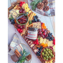 A festive charcuterie board with assorted cheeses, crackers, fruit, olives, nuts, and a 5 oz jar of Terrapin Ridge Farms Cranberry Relish With Grand Marnier™, garnished with herbs, pinecones, and a plaid napkin for cozy holiday entertaining.