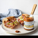 A sandwich with deli meat, lettuce, and onions is halved on a plate beside an open jar of Terrapin Ridge Farms Bleu Cheese Stuffed Olive Dip, a spreader in the jar. A blue towel appears in the background.