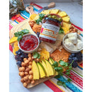 A vibrant charcuterie board features pineapple, cheese, crackers, chickpeas, mango, berries, mozzarella balls, cilantro, and a jar of Terrapin Ridge Farms Pineapple Aji Amarillo Jam on a colorful cloth.