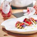 A chef delicately drizzles Taycte Acetaia Malpighi Prelibato Rosé Balsamic Vinegar—a pink Italian condimento known for its delicate and fruity flavor—over an artfully plated gourmet dish featuring colorful vegetables and fish on a pristine white plate. Holding both a spoon and the elegant Taycte bottle, the chef adds the final touch of this rose-hued dressing to complete the meal.