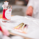 A bottle of Taycte Acetaia Malpighi Prelibato Rosé Balsamic Vinegar — a delicate and fruity pink Italian condimento — stands next to a clear plastic sheet covering sliced vegetables on the countertop, with a person blurred in the background.