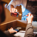 A person pours a dark liquid, Taycte Acetaia Malpighi Saba — an Italian cooked grape must syrup from Taycte, known as a traditional balsamic alternative from Modena Emilia-Romagna — from a glass bottle onto a spoon held by another person seated at a table. The scene suggests a tasting or sampling experience, with one individual wearing a striped apron.