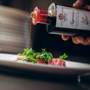 A hand pours Taycte Acetaia Malpighi Saporoso Balsamic Vinegar of Modena — a rich and savory Italian condiment from Taycte — from the bottle onto a dish with fresh greens and slices of meat, with a stack of plates visible in the softly blurred background.