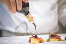 A person pours Taycte Acetaia Malpighi D.O.P. Traditional Balsamic Vinegar of Modena — Affinato Grade, aged 12+ years, from a bottle onto a gourmet dish featuring meat, potato, asparagus, pickled onion, and caviar on a white plate.
