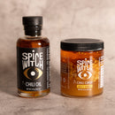 Spice Witch Chili Oil + Chili Crisp: two jars—chili oil with avocado oil and spicy honey chili crisp—feature black and gold labels with an eye graphic, all from Spice Witch, set against a neutral background.