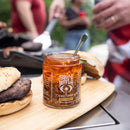 A box of 3 Spice Witch Chili Crisp jars from Spice Witch sits on a wooden board between burgers, while people grill and prepare food in the background.