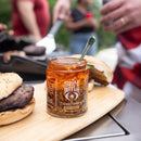 An open box of Spice Witch Chili Crisp Box of 2 sits on a wooden board next to burgers, while people grill and prepare food in the background.