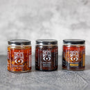 The Spice Witch Chili Crisp Box of 3, featuring original, black garlic, and spicy honey flavors, is displayed side by side on a gray textured surface with a neutral background.