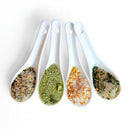 Four white ceramic spoons, each with a different SALTOPIA Infused Sea Salts blend from the Saltopia Ultimate Veggie Lover Salts set, are arranged in a row. The seasonings vary in color and texture, featuring green, orange, and mixed herbs.