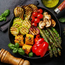 A plate of assorted grilled vegetables—like asparagus, corn, and zucchini—topped with fresh basil. SALTOPIA Infused Sea Salts’ Ultimate Veggie Lover Salts and a pepper mill boost flavor, with glistening olive oil nearby.
