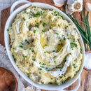 A white oval dish of creamy mashed potatoes topped with melted butter, chopped chives, and SALTOPIA Ultimate Veggie Lover Salts by SALTOPIA Infused Sea Salts. In the background are a wooden bowl, garlic, and fresh chives on a wooden surface.