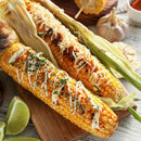 Two grilled corn cobs topped with cheese, herbs, and SALTOPIA Infused Sea Salts’ Ultimate Veggie Lover Salts, drizzled with sauce and served with lime wedges on a wooden board. Garlic and a sliced corn cob appear in the background.