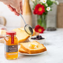 A hand drizzles Honey Truck Co Raw Florida Vanilla Infused Honey from a spoon onto buttered pound cake. Nearby sit a jar of Honey Truck Co honey, a bowl of berries, and a vase filled with red and white flowers atop a light marble surface.
