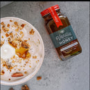 A bowl of yogurt topped with granola, nuts, seeds, and a drizzle of Honey Truck Co Raw Florida Vanilla Infused Honey sits next to a jar labeled Honey Truck Co on a gray surface. Some pieces of granola are scattered nearby.