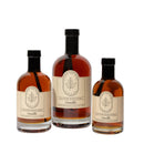Three glass bottles of Under The Bark Syrup Co Vanilla Maple Syrup, made with 100% pure maple syrup, feature beige labels with a maple leaf design and black caps, arranged together on a white background.