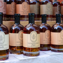 Bottles of Under The Bark Syrup Co Ginger Maple Syrup with beige labels reading “100% Pure Maple Syrup” are displayed on a table and shelves; this variety is infused with ginger root.