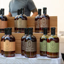 Bottles of Under The Bark Syrup Co Cardamom Maple Syrup are arranged on wooden shelves at a market stall, with a person behind the display showcasing these unique blends from Under The Bark Syrup Co.