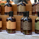 Bottles of Under The Bark Syrup Co Habanero Maple Syrup with decorative labels are neatly arranged on wooden shelves at a market stall.