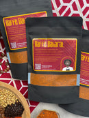 Three black bags of World Spice Kaffe Baharat are arranged on a geometric red and white cloth. Nearby sits a bowl filled with mixed whole spices, evoking the aromatic flavors of the Middle East. Each bag from World Spice features a chef illustration and text highlighting this unique coffee-infused blend.