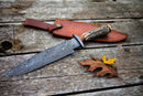 The Texan Knives Damascus Steel 1800s Bowie Knife, 15" with a Damascus steel guard and stag horn handle, sits on weathered wood next to its brown leather sheath and two fallen leaves.