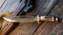 A Texan Knives Damascus Steel Vintage Bowie Knife – 13" with brass guard and stag horn handle, comes with a leather sheath, is displayed on wood beside a rifle with a leather strap.