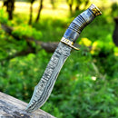 A Texan Knives Vintage Damascus Steel Bowie Knife for sale—13" with a brass guard and camel bone handle—stands upright in a log, its patterned blade gleaming against a blurred green forest backdrop.