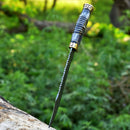 The Texan Knives Vintage Original Damascus Steel Bowie Knife – 13" with a brass guard and camel bone handle is stabbed upright into a tree trunk, with blurred green foliage in the background.
