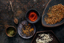 A selection of spices is arranged on dark dishes—mustard seeds, chunky paprika, dried herbs, and crushed seasonings—along with a spoon holding World Spice Voodoo by World Spice, all set against a textured dark background.