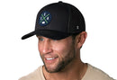 Smiling man with stubble wearing a Haka Hat Seattle Trucker Hat | Black SEA X Snapback with a blue and green circular SEA emblem and a beige t-shirt, set against a plain white background.