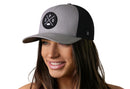 A woman with long brown hair smiles while wearing the Haka Hat Spokane Trucker Hat in gray and black, featuring a round black logo. This comfortable snapback cap by Haka Hat is shown against a white background.