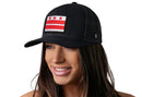 A woman with long brown hair smiles while wearing the Haka Hat Washington DC Flag Trucker Hat | Black Snapback, featuring an embroidered DC flag patch with three red stars and two red bars. Plain white background. Brand: Haka Hat.
