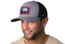 A smiling man with a trimmed beard wears the Haka Hat Wyoming Flag Trucker Hat, Gray-Black WY Snapback, featuring a white bison logo in a red-bordered patch. He has light eyes, a beige shirt, and stands against a white background.