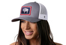 A woman with long brown hair smiles while wearing the Haka Hat Wyoming Flag Trucker Hat, a gray and white snapback featuring a patch of a white buffalo silhouette inside a red and blue rectangular border.