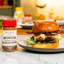 A cheeseburger topped with bacon, mushrooms, and greens sits on a plate next to a jar of Powerful Pepper Company Cajun Blackening Seasoning on a marble countertop, delivering the bold flavors of Cajun blackened cuisine to your kitchen.