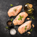 Three boneless skinless chicken breasts from the Dude Food Dude Meat Box, seasoned with herbs, peppercorns, sea salt, and garlic cloves, alongside a bottle of olive oil on a dark surface.