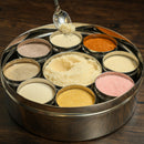 A round metal Spice Wheel Flavored Sugar Spice Wheel by Spice Wheel, featuring nine sections each filled with a distinctively colored flavored sugar. A spoon lifts a yellowish sugar from the center compartment, while the elegant wheel rests on a dark wooden surface.