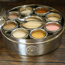 The Spice Wheel Flavored Sugar Spice Wheel by Spice Wheel features a round metal design with nine compartments, each containing a different flavored sugar powder. It includes a small metal spoon and has a partially open lid, displayed on a wooden surface.