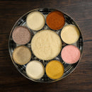 The Spice Wheel Flavored Sugar Spice Wheel by Spice Wheel is a round metal spice box, designed in the style of a traditional spice wheel, with nine compartments each filled with a unique flavored sugar or seasoning, elegantly displayed on a dark wooden surface.
