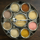 The Spice Wheel Flavored Sugar Spice Wheel by Spice Wheel features eight small round metal containers filled with a variety of flavored sugars and spices, arranged in a circular metal wheel with a spoon resting on the center container, all displayed on a dark wooden surface.