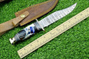 The Texan Knives Hand-Forged Western Boulder Colorado Knife features an 11" Damascus steel blade, brass guard, and resin grip. It rests by a brown leather sheath and wooden ruler on green artificial grass.