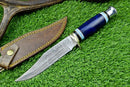 The Texan Knives Western Boulder Colorado Knife features an 11" handmade Damascus steel blade with intricate patterns, a blue epoxy resin handle, and comes with a brown leather sheath with yellow stitching and snap closure.