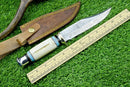 The Texan Knives Hand-Forged Western Boulder Colorado Knife features an 11” razor-sharp Damascus blade, a sleek white resin grip, and decorative accents. It comes with a brown leather sheath and is displayed beside a wooden ruler on green artificial grass.