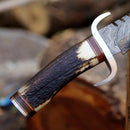 Close-up of the Texan Knives Damascus Steel Western Bowie Knife – 12" with a patterned blade, stag horn handle, and Damascus steel guard, shown against a blurred natural background. Includes leather sheath.