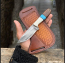 A hand holds the Texan Knives Western Knife, featuring a patterned Damascus steel blade, resin handle with blue accent, and leather belt loop sheath. Stone path and walls are seen in the background.